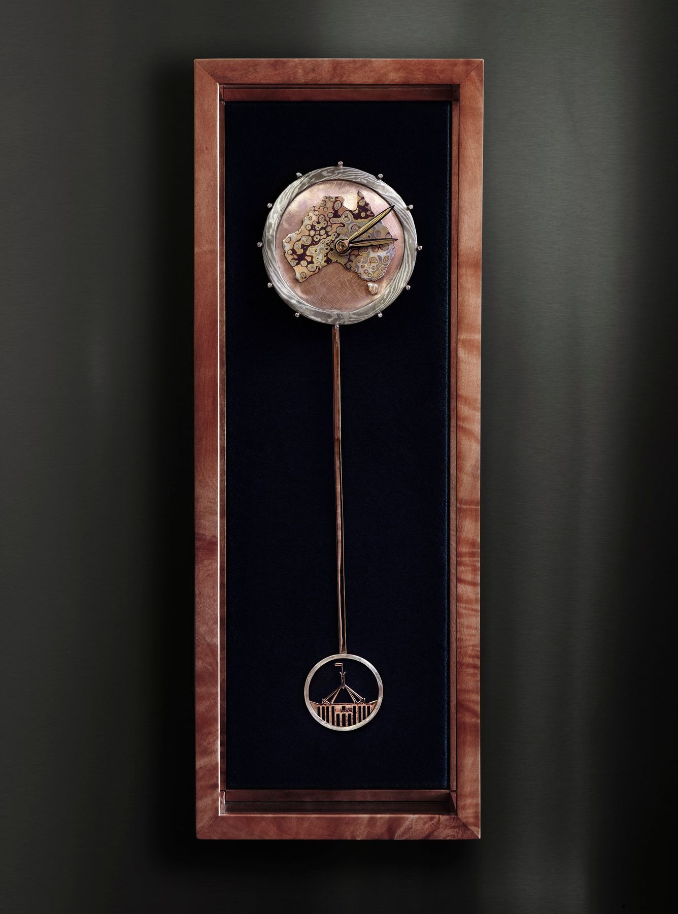 The Always Clock in its Tasmanian myrtle case: the Mokume Gane face above a long rose gold pendulum ending in a ring framing Parliament House