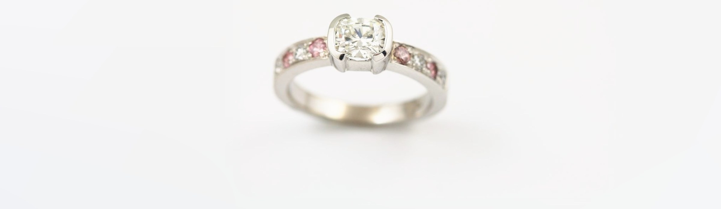 A cushion-cut diamond in a white gold half-bezel, with pink and white diamonds set along the band