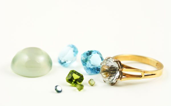 Loose stones and a worn gold ring laid out on white: a green cabochon, blue topaz, peridot and a small diamond